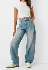 Světle modré široké džíny vyrobené z denimové látky, s vysokým pasem, předními kapsami a vybledlým vzhledem. Kombinované s bílou zkrácenou košilí a teniskami.