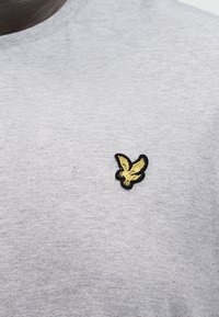 Κεντητός κίτρινος αετός με μαύρο περίγραμμα στο στήθος ενός πουκάμισου από ανοιχτό γκρι ύφασμα.