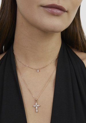 Close-up of a woman wearing two gold necklaces: a small pendant and a larger cross-shaped pendant with clear gemstones.