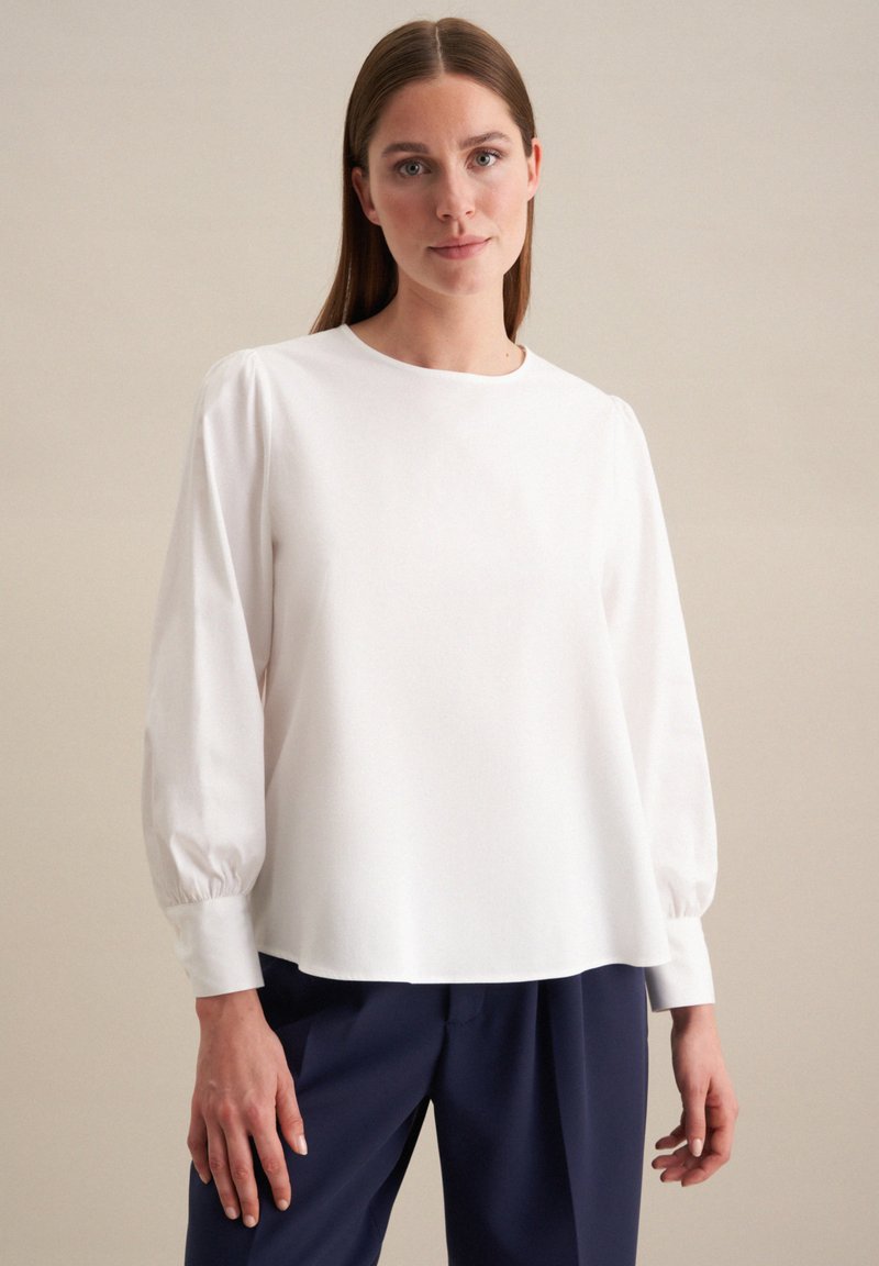 Blusa bianca a maniche lunghe con scollo rotondo, maniche arricciate e vestibilità rilassata. Il tessuto sembra liscio e leggero.