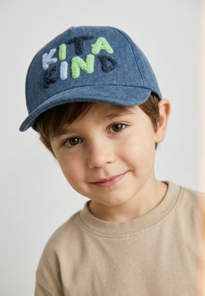 Young boy wearing a blue cap with "Kinda Kind" embroidered in multicolored letters and a beige crewneck shirt, smiling gently.