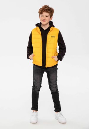Gilet rembourré jaune sur un sweat à capuche noir, avec une fermeture éclair et deux poches avant. Porté avec un jean foncé et des baskets blanches, se tenant devant un fond uni.