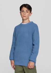 Niebieski sweter z dzianiny z prążkowanym dekoltem i mankietami, z fakturowanym wzorem. Noszony na białej koszuli, w zestawieniu z oliwkowymi spodniami.