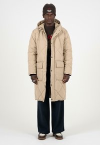 Beige gewatteerde lange jas met een capuchon, voorzien van drukknoopsluitingen en twee voorzakken. Gedragen over een donkere trui en brede broek.