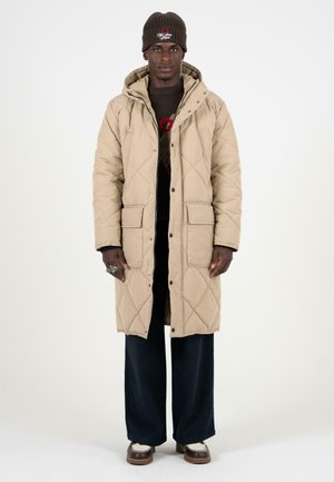 Manteau long beige matelassé avec capuche, doté de fermetures à pression et de deux poches avant. Porté par-dessus un pull sombre et un pantalon large.