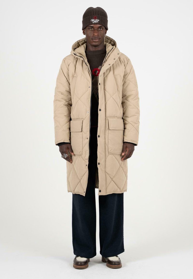Beige gewatteerde lange jas met een capuchon, voorzien van drukknoopsluitingen en twee voorzakken. Gedragen over een donkere trui en brede broek.