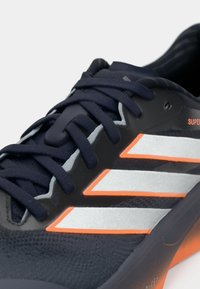 Niebieski but sportowy z teksturowaną siateczkową cholewką, trzema srebrnymi paskami, pomarańczowymi akcentami i wzorzystymi sznurowadłami oraz wyściełanym językiem.