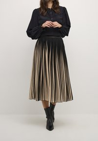 Woman wearing a black long-sleeve blouse with buttons and a pleated midi skirt with black and beige vertical stripes, paired with black ankle boots.