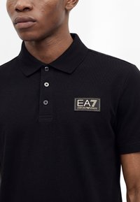 Jeune homme portant un polo noir EA7 Emporio Armani avec trois boutons et un écusson logo sur la poitrine gauche.