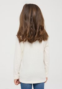 Camicia a maniche lunghe di colore crema realizzata in tessuto morbido, con scollatura rotonda e orlo leggermente curvo, abbinata a jeans in denim blu.