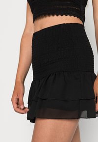 Saia mini preta com pregas, com bainha em camadas. Textura suave, cintura elástica, material leve e detalhes transparentes na bainha.