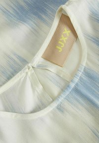 Lätt tygtröja i en blandning av vita och mjuka blå streck, med rund halsringning och en etikett som visar "JUXX" i gult.