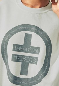 Maglietta bianca con grafica circolare raffigurante un segno più, la scritta "prendi quello 1993" e nomi di città lungo il bordo.