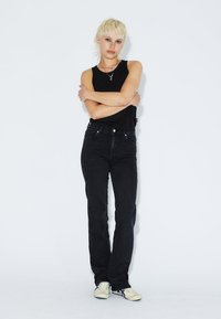 Schwarze High-Waist-Jeans mit geradem Bein und Fronttaschen, kombiniert mit einem schwarzen ärmellosen Top und hellen Sneakers.