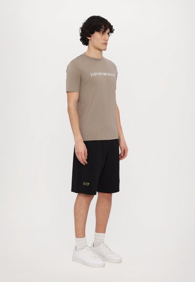 Brown cotton T-shirt with "Emporio Armani" text, paired with black shorts featuring gold “EA7” logo. Model wears white sneakers and socks.