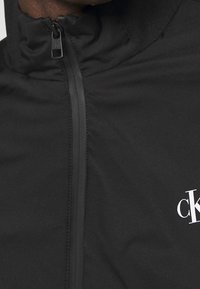 Giacca leggera nera con colletto alto, zip frontale e logo bianco sul lato sinistro. Texture liscia e design minimalista.