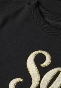Sweat-shirt noir avec col côtelé présentant une lettre S blanche texturée brodée, soulignée de fil jaune.