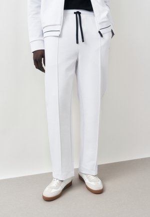 Muž nosiace biele nohavice na šnúrky s čiernymi šnúrkami, bielu bundu na zips a biele tenisky s hnedými podrážkami stojaci na šedej podlahe.