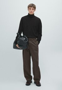Il modello indossa un maglione a collo alto nero e pantaloni larghi marroni, tenendo una grande borsa in pelle nera con accenti hardware a contrasto.