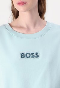 Světle modrá mikina s kulatým výstřihem a žebrovaným okrajem. Na hrudi se nachází výrazné logo "BOSS" s texturovanými modrými detaily.