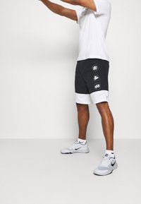 Pantalones cortos deportivos en blanco y negro con patrones geométricos y logotipos, fabricados en tela ligera. Modelo combinado con zapatillas Nike blancas.