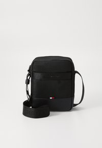 Black crossbody bag made of fabric with a textured finish. Smooth synthetic bottom, adjustable strap, and logo detail with red, white, and blue accents.