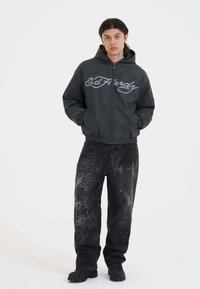 Una giovane persona indossa una felpa nera con zip Ed Hardy e jeans neri con stampe grafiche scure davanti a uno sfondo bianco uniforme.