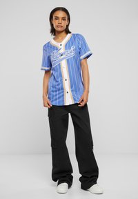 Maglietta blu a maniche corte con righe bianche e beige, dotata di un'apertura frontale e dettagli testuali grafici. Abbinata a pantaloni cargo neri.