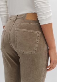 Béžové slim-fit kalhoty vyrobené z měkké látky, s kapsami na zadní straně a texturovanou koženou nášivkou na pase.