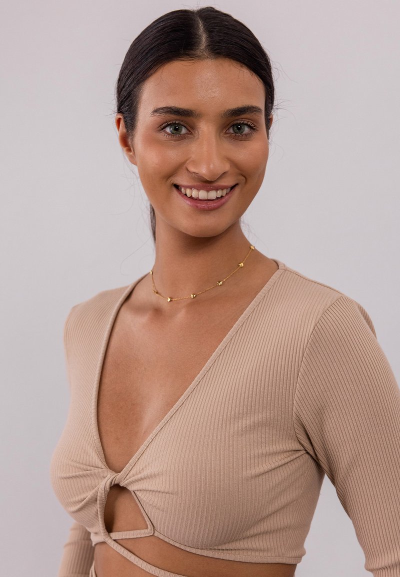 Beige gestreifter Langarm-Top mit tiefem V-Ausschnitt und Knotendetail vorne sowie Ausschnitt. Getragen mit einer zarten Goldkette.