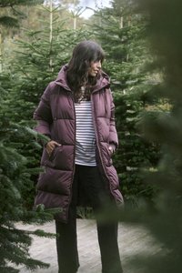 Lungo cappotto puffa viola con cappuccio, caratterizzato da sezioni trapuntate e tasche laterali. Indossato sopra una maglietta a righe, ambientato tra alberi sempreverdi.