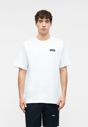 Junger Mann mit kurzen lockigen Haaren, der ein weißes T-Shirt mit kurzen Ärmeln und schwarze Shorts trägt, steht vor einem schlichten hellen Hintergrund.