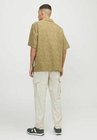 Camisa de manga corta de color verde oliva con estampado, combinada con pantalones cargo de color beige claro. Presenta un corte holgado y un diseño de cuello abierto.