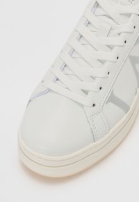 Sneaker en cuir blanc avec une texture lisse, lacets blancs plats et un logo discret sur le côté. Bout arrondi et semelle en caoutchouc blanc cassé.