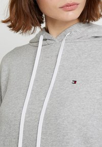 Felpa grigia realizzata in tessuto morbido. Presenta un cappuccio con coulisse e un piccolo logo ricamato sul petto in rosso, bianco e blu.