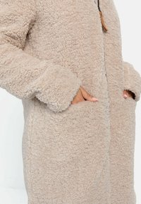 Manteau en polaire beige avec un tissu doux et texturé, doté de poches latérales et d'une fermeture éclair à l'avant, coupe légèrement ample et sans attaches visibles.