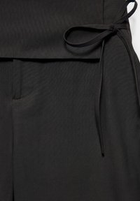 Pantalon noir avec un nœud en tissu à la taille sur le côté droit et une petite poche sur le devant gauche.