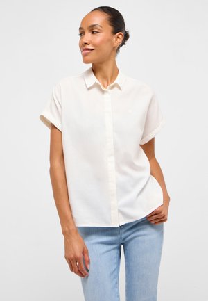 Frau mit dunklem Haar, die ein weißes Kurzarmhemd mit Knöpfen und hellblaue Jeans trägt, steht mit einer Hand in der Tasche und schaut seitlich.