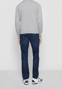 Uomo che indossa una camicia a maniche lunghe grigia, jeans blu scuro e sneakers bianche, in piedi, visto da dietro su uno sfondo chiaro e uniforme.