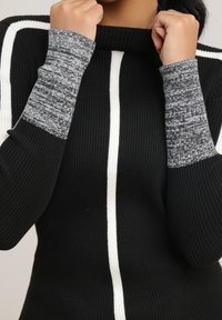 Personne portant un pull noir côtelé avec des rayures verticales blanches et des poignets gris mouchetés, tenant le col près de son cou.