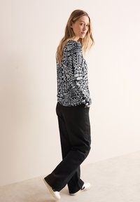 Haut à manches longues noir et blanc à motifs géométriques, associé à un pantalon noir et des baskets blanches, se tenant devant un fond clair.