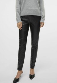Pantalons en cuir synthétique noir à coupe slim et texture lisse. Portés avec un pull gris et des escarpins noirs à bout pointu.