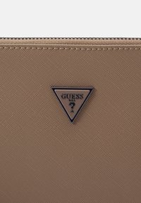 Portfel z teksturowanej tkaniny w kolorze wielbłądzim z czarnym metalowym logo w kształcie trójkąta. Posiada zamek błyskawiczny oraz widoczne przeszycia wzdłuż brzegów.