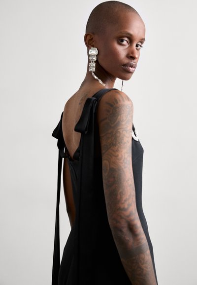 Vestido negro con un diseño sin espalda, detalles en forma de lazo en los hombros y un acento plateado en el frente. Pendientes grandes y ornamentados con patrones intrincados.