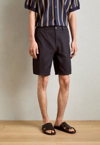 Sorte shorts med en klassisk pasform, kombinert med en kortermet stripete marineblå skjorte. Person iført brune tøfler, står på tregulv.