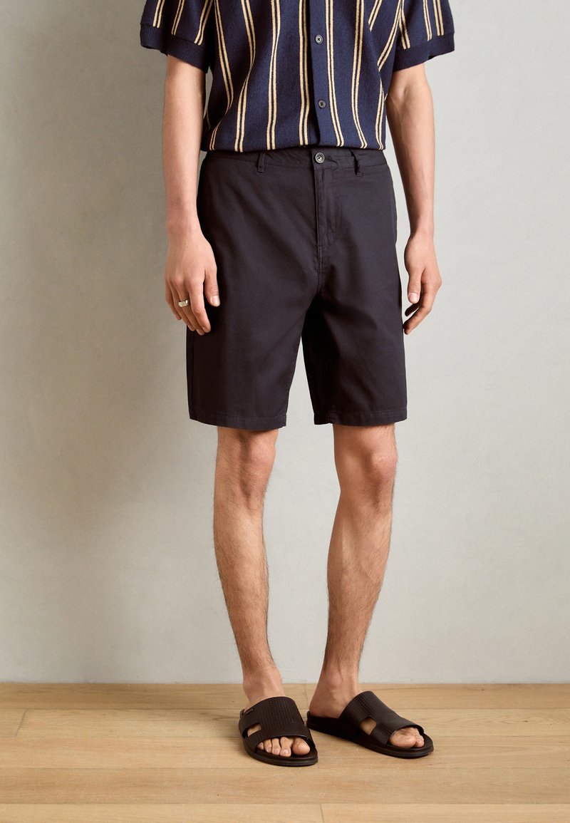 Sorte shorts med en klassisk pasform, kombinert med en kortermet stripete marineblå skjorte. Person iført brune tøfler, står på tregulv.