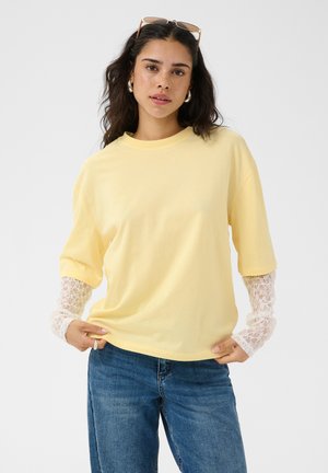 Jeune femme aux cheveux ondulés et foncés porte des lunettes de soleil rondes, un haut jaune à manches courtes superposé à des manches en dentelle blanche, et un jean bleu.