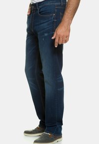 Jeans de mezclilla azul oscuro con un corte slim, que presentan un ligero desgaste, costuras naranjas, bolsillos frontales y una bragueta con cremallera. Combinados con zapatos grises.