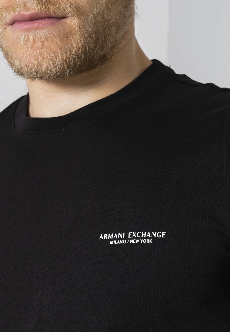 Homme avec une légère barbe portant un t-shirt noir Armani Exchange avec un texte blanc indiquant Milano / New York sur la poitrine.