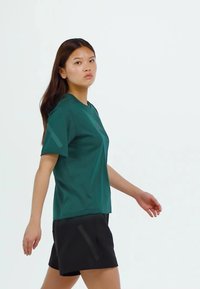 Zelené tričko s krátkym rukávom vyrobené z hladkej látky, nosené s čiernymi šortkami. Tričko má okrúhly výstrih a voľný strih.